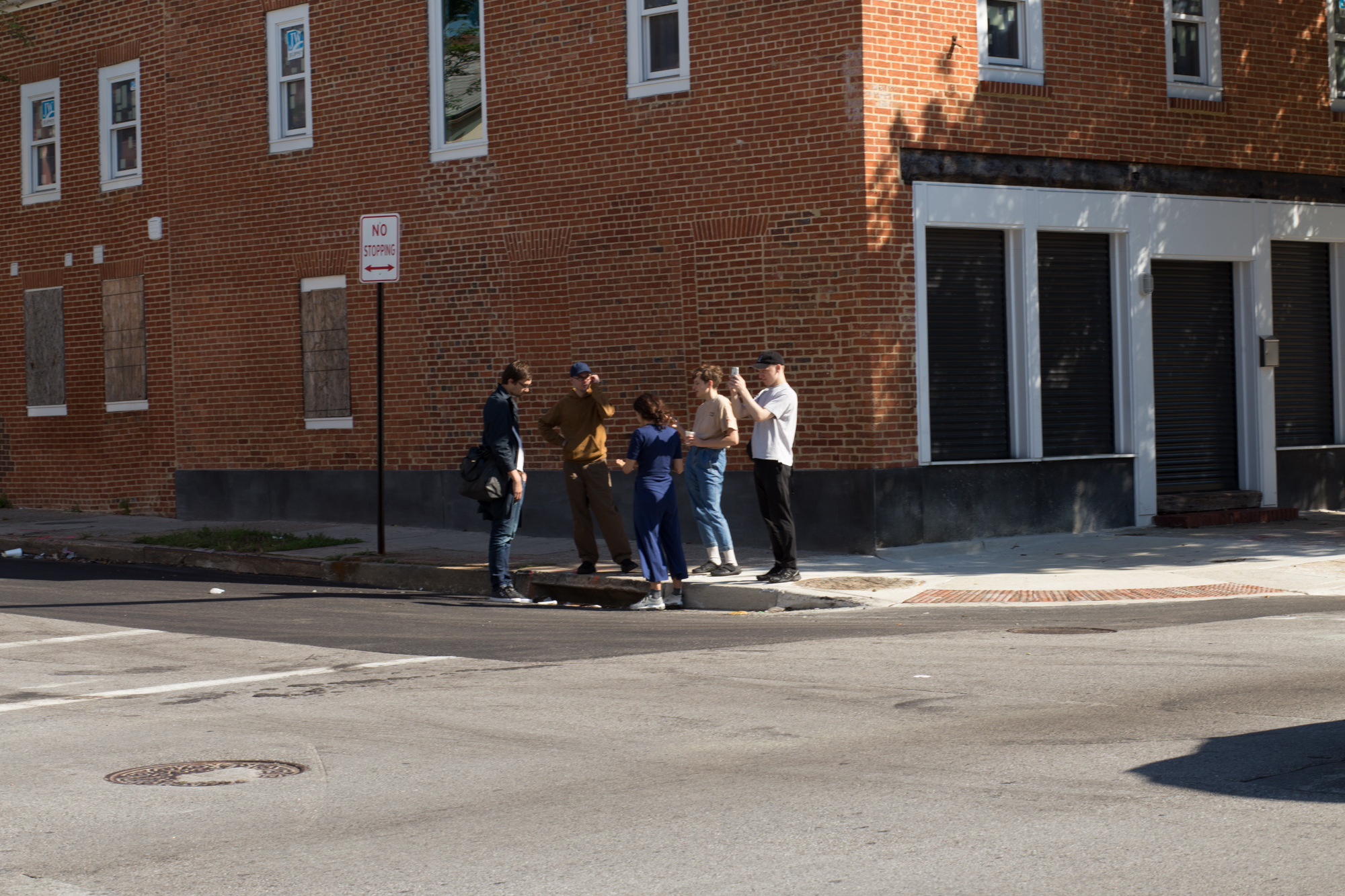 Five people stand on a street corner in front of a brick building; one takes a photo with his phone