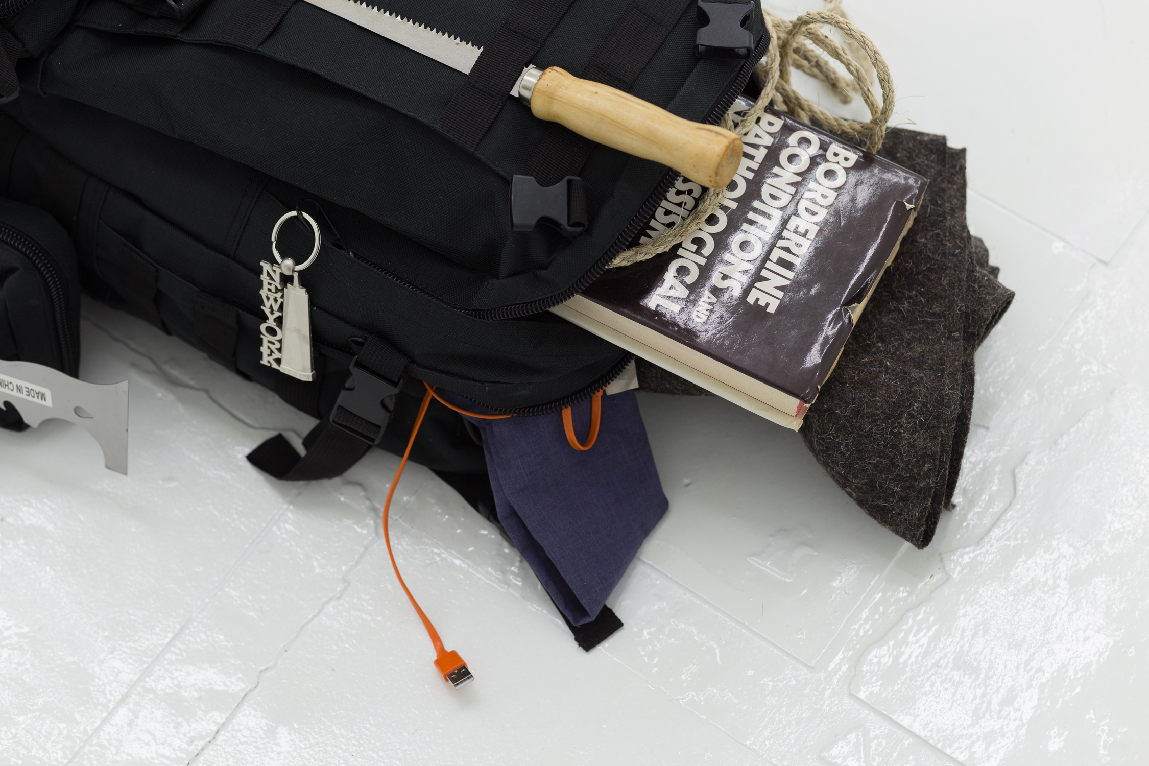  Some items stick out of the backpack, which has a New York keychain on its zipper. A book, a drywall knife, a usb cable, and a piece of felt